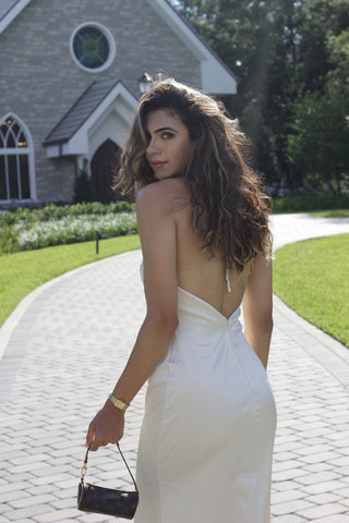 Model wearing a cream, satin, halter dress, that ties around neck. The model is showing the back of the dress which is a low back. Model is standing in front of a church.