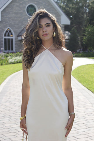 Model wearing a cream, satin, halter dress, that ties around neck. Model is standing in front of a chapel. 