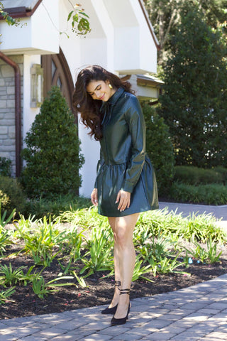 Model is wearing deep green vegan leather dress with drop waist, high collar and long sleeves. Model is standing in front of church and greenery. 