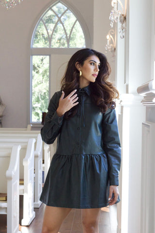 Model is wearing deep green vegan leather dress with drop waist, high collar and long sleeves. Model is standing in a white church with beautiful windows in the back.