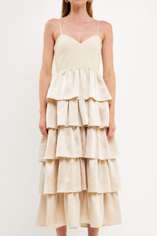 Model wearing champagne color dress with tiered ruffles on the skirt. The top is a spaghetti strap, sweetheart neckline bust.Model is standing in front of a white background and the image shows her whole body. The image is up close to show the detail of the dress. 