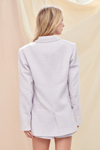 Model is standing in front of a cream backdrop. She is wearing a lavender and white plaid and tweed blazer and short set. Image is showing the back of the blazer. 