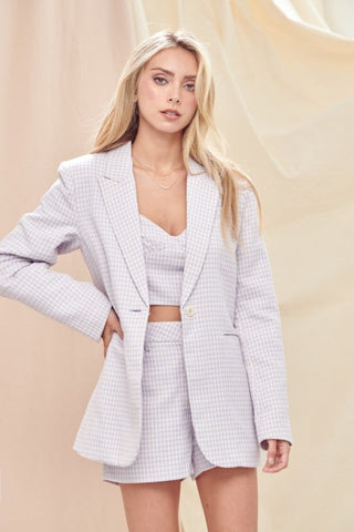 Model is standing in front of a cream backdrop. She is wearing a lavender and white plaid and tweed blazer and short set. Model is looking at the camera.  Model has one hand on her hip.