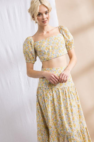 Model standing in front of white and tan backdrop. Wearing a flower patterned, two-piece, skirt set. Sleeves are puffy. Model is facing the front.