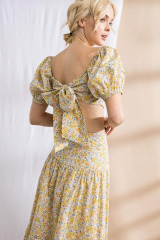 Model standing in front of white and tan backdrop. Wearing a flower patterned, two-piece, skirt set. Sleeves are puffy. Model is showing the bow in the back that ties the shirt together.