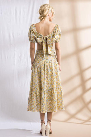 Model standing in front of white and tan backdrop. Wearing a flower patterned, two-piece, skirt set. Sleeves are puffy. Model is facing to the back and showing the large bow that ties the top.