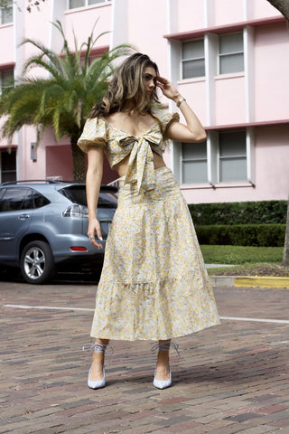 Model standing in front of pink building. Wearing a flower patterned, two-piece, skirt set. Sleeves are puffy and bow is tied in the front.