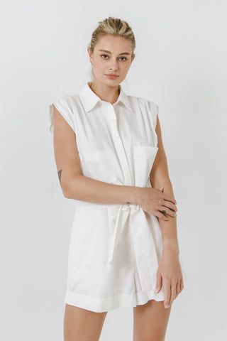 Model wearing white, collared, cotton, belted romper. She is standing in front of a white backdrop and is looking into the camera. The front of the romper is showing. 