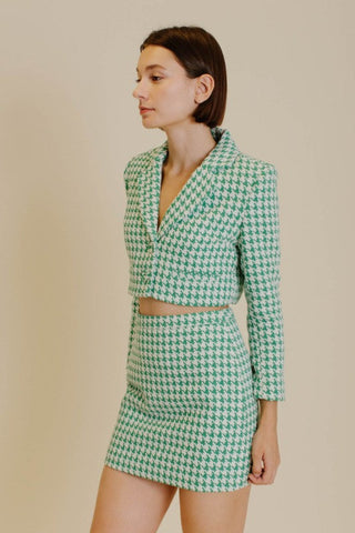 Model wearing a blazer and skirt set that is a emerald green and white houndstooth tweed. The model is standing in front of a tan background and showing the front of the set. 