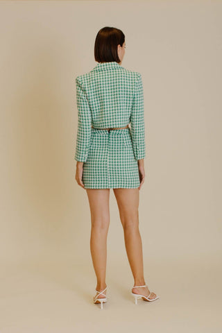 Model wearing a blazer and skirt set that is a emerald green and white houndstooth tweed. The model is standing in front of a tan background and showing the back of the set. 