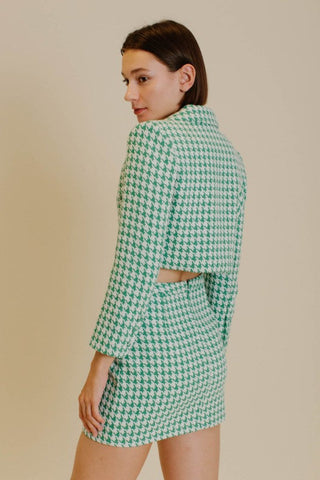Model wearing a blazer and skirt set that is a emerald green and white houndstooth tweed. The model is standing in front of a tan background and showing the back of the set. 