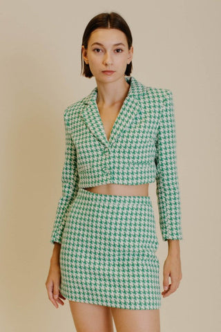 Model wearing a blazer and skirt set that is a emerald green and white houndstooth tweed. The model is standing in front of a tan background and showing the front of the set. 