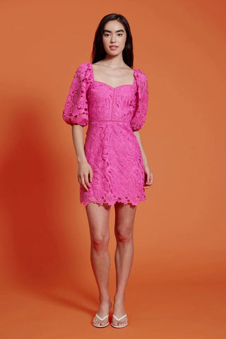Model standing in front of orange background. She is wearing a lace pink dress with puff sleeves. The Dress has laser cutouts.  