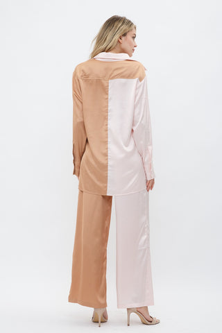 Model is standing in front of white backdrop. She is facing backwards to show the back of the outfit. She is wearing a satin long sleeve top and satin pant set. The top is a color block satin top with one side peach and one side light pink. The pants are a color block satin pant with one side peach and the other side light pink. 