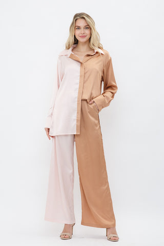 Model is standing in front of white backdrop. She has one hand in her pocket and the other by her side. She is wearing a satin long sleeve top and satin pant set. The top is a color block satin top with one side peach and one side light pink. The pants are a color block satin pant with one side peach and the other side light pink. 