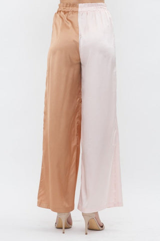 Model is standing in front of white backdrop. She is wearing a satin long sleeve top and satin pant set. The top is a color block satin top with one side peach and one side light pink. The pants are a color block satin pant with one side peach and the other side light pink.  The photo is of the pants from the back.