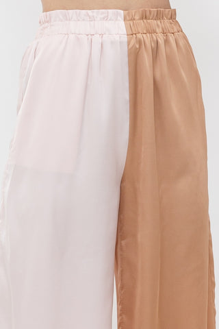 Model is standing in front of white backdrop. She is wearing a satin long sleeve top and satin pant set. The top is a color block satin top with one side peach and one side light pink. The pants are a color block satin pant with one side peach and the other side light pink. Image is a close up of the pants.