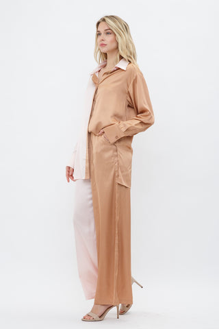 Model is standing in front of white backdrop. She has one hand in her pocket and the other by her side. She is facing slightly diagonal. She is wearing a satin long sleeve top and satin pant set. The top is a color block satin top with one side peach and one side light pink. The pants are a color block satin pant with one side peach and the other side light pink. 