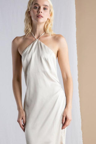 Model wearing a cream, satin, halter dress, that ties around neck. Model is standing in front of a tan and white background.