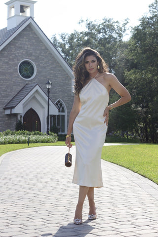 Model wearing a cream, satin, halter dress, that ties around neck. Model is standing in front of church. 
