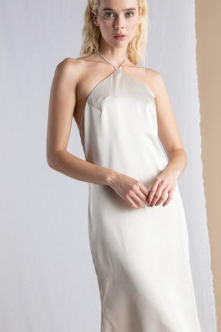Model wearing a cream, satin, halter dress, that ties around neck. Model is standing in front of a tan and white background.