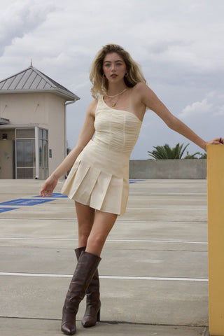 Model wearing cream, pleated, tennis skirt. She is standing in parking garage on yellow lift. She paired the cream skirt with brown boots and cream bodysuit.