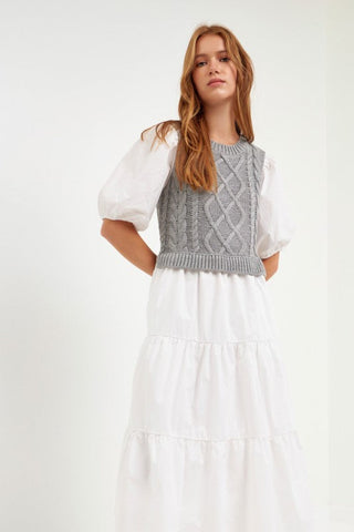 Model wearing white dress with grey cable knit sweater over the dress. Model is standing in front of white backdrop. 