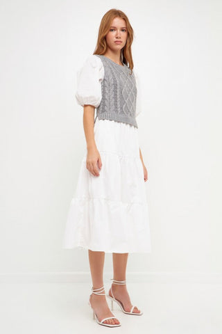 Model wearing white dress with grey cable knit sweater over the dress. Model is standing in front of white backdrop. 