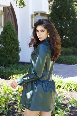 Model is wearing deep green vegan leather dress with drop waist, high collar and long sleeves. Model is standing in front of church with greenery in the front.