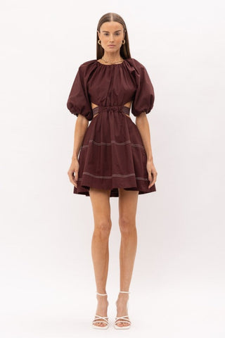 Brown cut out midi dress with white thread detailing, puff sleeves, and a bow enclosure. Model is standing in front of white background.