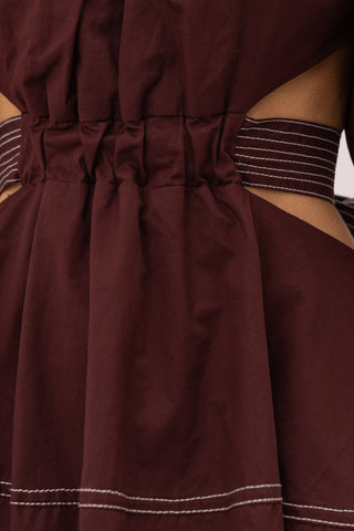 Brown cut out midi dress with white thread detailing, puff sleeves, and a bow enclosure. Model is standing in front of white background. The image is close up to show the detail of the dress.