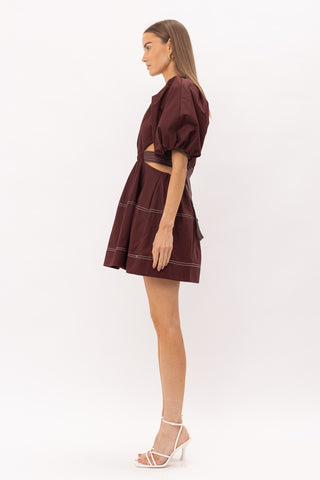 Brown cut out midi dress with white thread detailing, puff sleeves, and a bow enclosure. Model is standing in front of white background. Photo is taken of model facing sideways.