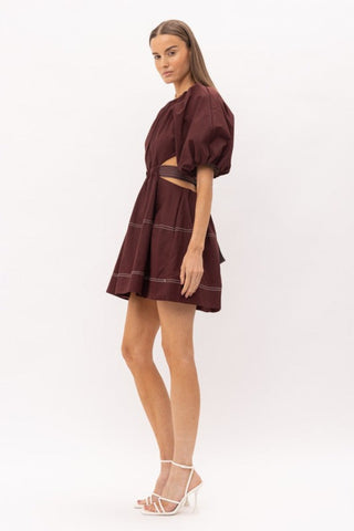 Brown cut out midi dress with white thread detailing, puff sleeves, and a bow enclosure. Model is standing in front of white background. Model is facing the side to show the side of the dress.