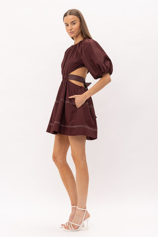 Brown cut out midi dress with white thread detailing, puff sleeves, and a bow enclosure. Model is standing in front of white background. Model is standing side and showing the pocket detail.