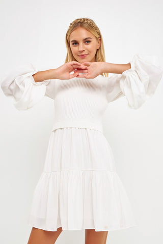 Model is wearing a white dress with a white mock neck knit vest over it. Model is standing in front of white background.  