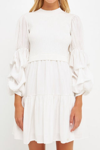 Model is wearing a white dress with a white mock neck knit vest over it. Model is standing in front of white background.  Photo is showing the dress up close.
