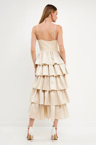 Model wearing champagne color dress with tiered ruffles on the skirt. The top is a spaghetti strap, sweetheart neckline bust.Model is standing in front of a white background and the image shows her whole body. Model is turned around to show the back of the dress. 
