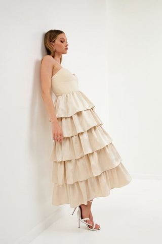 Model wearing champagne color dress with tiered ruffles on the skirt. The top is a spaghetti strap, sweetheart neckline bust.Model is standing in front of a white background and the image shows her whole body. Model is leaning up against wall