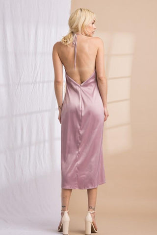 Model wearing a purple, mauve, satin, halter, midi dress. Model standing in front of a backdrop that is half white and half tan. The image is showing the dress from behind.