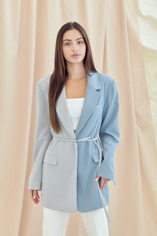 Model standing in front of a cream draped curtain. Model is wearing white pants and a white cropped top with a blue oversized color-block blazer. Blazer colors are light blue on the left side and dark blue on the right side. It has a thin belt that belts the blazer in the center. Photo is up close and model is looking right at the camera.  