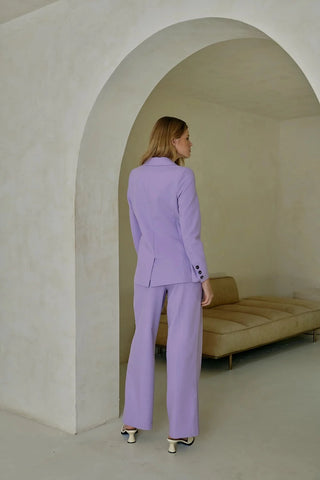 Model is standing in front of an archway. Model is wearing purple, pleated pant with matching purple blazer. Model is facing back to show the front of the pants. 