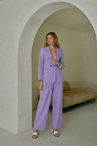 Model is standing in front of an archway. Model is wearing purple, pleated pant with matching purple blazer. Model is facing forward to show the front of the pants. 