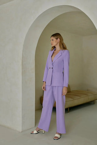 Model is standing in front of an archway. Model is wearing purple, pleated pant with matching purple blazer. Model is facing to the side to show the front of the pants. 