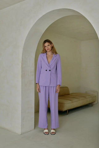 Model is standing in front of an archway. Model is wearing purple, pleated pant with matching purple blazer. Model is facing forward to show the front of the pants. 