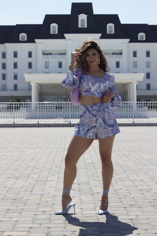 Model wearing paisley, pastel short set. She is standing in front of a large white building. She is posed facing forward with blue heels and a purple purse 