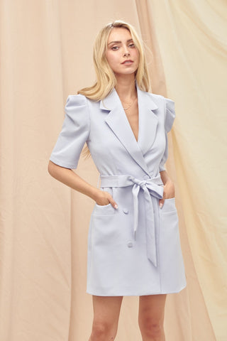 Model standing in front of cream draped background. She is wearing a purple blazer dress with puffed sleeves. She has her hands in her pockets and is looking directly at the camera. 