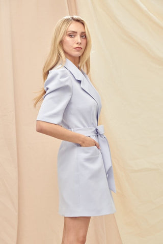 Model standing in front of cream draped background. She is wearing a purple blazer dress with puffed sleeves. She has her hands in her pockets and is facing sideways to show the side of the dress.