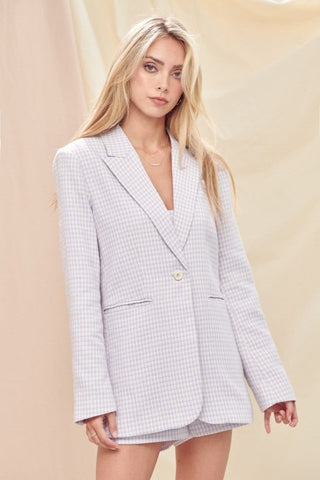 Model is standing in front of a cream backdrop. She is wearing a lavender and white plaid and tweed blazer and short set. Model is looking at the camera.  