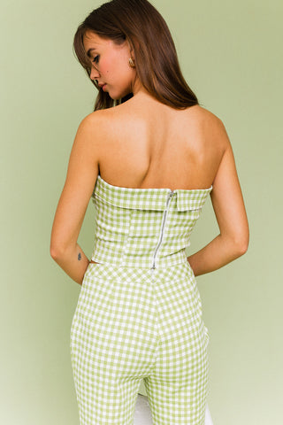 Model wearing a green and white gingham matching set. The top is a gingham corset, tube top. The pants are a straight leg, cropped gingham pant.  Model is standing in front of green background.  Photo of back of corset. 