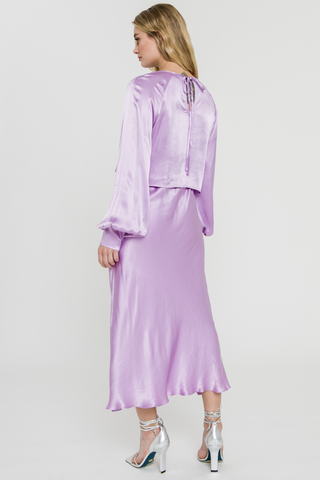 Model wearing a purple, satin, midi, skirt. She is standing in front of a white backdrop and wearing a matching top. She is facing the back to show the back of the skirt. 